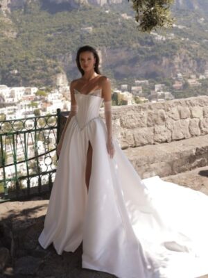 A bride shows a full view of the front of her Micol by Eden Aharon wedding dress. This strapless dress features a corset bodice that is adorned with a dropped Basque waistline. It’s made of silk Mikado fabric and features a slit. See it in Chicago, IL.