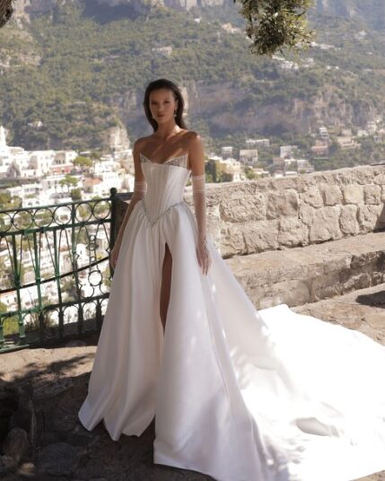 A bride shows a full view of the front of her Micol by Eden Aharon wedding dress. This strapless dress features a corset bodice that is adorned with a dropped Basque waistline. It’s made of silk Mikado fabric and features a slit. See it in Chicago, IL.