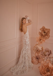 A bride shows the dramatic train on the back of her dreamy wedding dress, Lilac by Eisen Stein. The high neckline halter bridal gown with floral lace sleeves features a fit and flare silhouette made of lace with 3D floral texture. See it at a bridal shop in Chicago, IL.