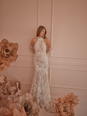 A bride shows the front of her dreamy wedding dress, Lilac by Eisen Stein. The high neckline halter bridal gown with floral lace sleeves features a fit and flare silhouette made of lace with 3D floral texture. See it at a bridal shop in Chicago, IL.