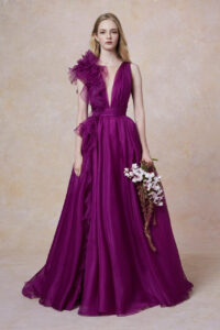 A bridesmaid holds flowers and shows her bridesmaid's dress, 32821 by Marchesa. The stunning purple evening gown has a plunging neckline, a cascading ruffle, and a banded waist. It is available at a boutique in Chicago, IL.