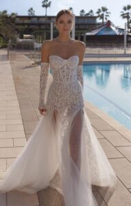 A bride steps forward and shows the front of her wedding dress, Natty by Eden Aharon. The mermaid bridal gown features a sweetheart neckline, lace detailing, and a train. Available in Chicago, IL.