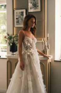 A bride stands in front of an end table and shows the side detailing on the Sheila by Neta Dover wedding dress. This A-line bridal gown features thin straps, flora embellishments on the sides of the waist and down the skirt, a corset bodice with exposed boning, and a dramatic train. Available at a bridal shop in Chicago, IL.