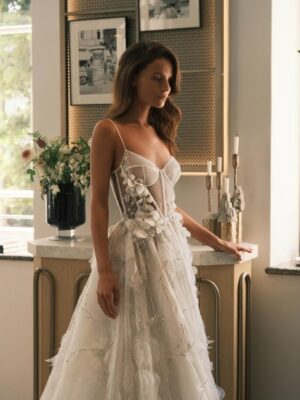 A bride stands in front of an end table and shows the side detailing on the Sheila by Neta Dover wedding dress. This A-line bridal gown features thin straps, flora embellishments on the sides of the waist and down the skirt, a corset bodice with exposed boning, and a dramatic train. Available at a bridal shop in Chicago, IL.