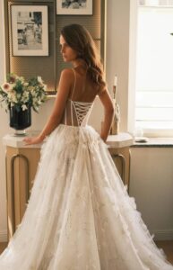 A bride stands in front of an end table and shows the back detailing of the Sheila by Neta Dover wedding dress. This A-line bridal gown features thin straps, flora embellishments on the sides of the waist and down the skirt, a corset bodice with exposed boning, and a dramatic train. Available at a bridal shop in Chicago, IL.