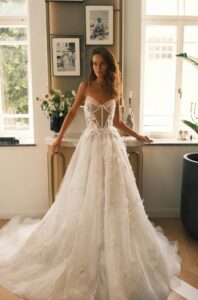 A bride stands in front of an end table and shows the Sheila by Neta Dover wedding dress. This A-line bridal gown features thin straps, flora embellishments on the sides of the waist and down the skirt, a corset bodice with exposed boning, and a dramatic train. Available at a bridal shop in Chicago, IL.