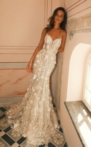 A bride wearing the Stephanie wedding dress by Neta Dover leans against a wall. The bridal gown features a fitted silhouette made of silk tulle with a layer of floral lace. This gown features delicate 3D floral embellishments, thin straps, a belt detail, and a sweetheart neckline. See it at a bridal shop in Chicago, IL.