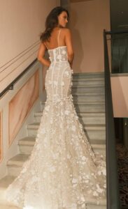 A bride shows the back of her wedding dress, Stephanie by Neta Dover. The bridal gown features a fitted silhouette made of silk tulle with a layer of floral lace. It has delicate 3D floral embellishments, thin straps, a belt detail, and a train. See it at a bridal shop in Chicago, IL.