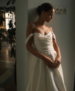 A close-up image of a bride lowering her eyes while wearing the Sydney by Neta Dover wedding dress. This elegant bridal gown features a pleated crossover bodice, a long pleated skirt completed, and off-the-shoulder sleeves. See it at a bridal shop in Chicago, IL.