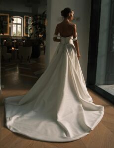 A rear view of a bride showing the dramatic train in the Sydney by Neta Dover wedding dress. This elegant wedding bridal gown features a pleated crossover bodice, a long pleated skirt, and off-the-shoulder sleeves. See it at a bridal shop in Chicago, IL.