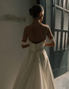 A rear view of a bride showing the back of the Sydney by Neta Dover wedding dress. This elegant wedding bridal gown features a pleated crossover bodice, a long pleated skirt, and off-the-shoulder sleeves. Available at a bridal shop in Chicago, IL.