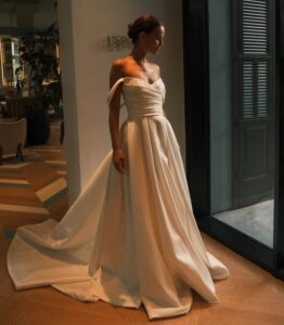 A bride lowers her eyes while showing the front of the Sydney by Neta Dover wedding dress. This elegant wedding bridal gown features a pleated crossover bodice, a long pleated skirt with a train, and off-the-shoulder sleeves. See it at a bridal shop in Chicago, IL.