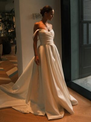 A bride lowers her eyes while showing the front of the Sydney by Neta Dover wedding dress. This elegant wedding bridal gown features a pleated crossover bodice, a long pleated skirt with a train, and off-the-shoulder sleeves. See it at a bridal shop in Chicago, IL.