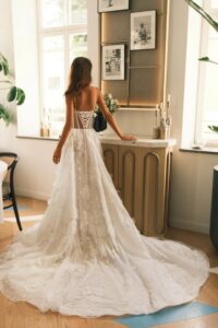 A bride shows the back of the Sheila by Neta Dover wedding dress. This A-line bridal gown features thin straps, flora embellishments on the sides of the waist and down the skirt, a corset bodice with exposed boning, and a dramatic train. See it at a bridal shop in Chicago, IL.