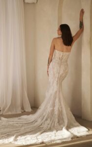 A bride shows the back of her 1752 wedding dress by Martina Liana. It is a lace bridal gown with a sweetheart neckline, sheer strapless bodice, and dramatic train with buttons. The fit and flare silhouette is shown without the detachable off-the-shoulder long lace sleeves. It is available at a bridal shop in Chicago, IL.