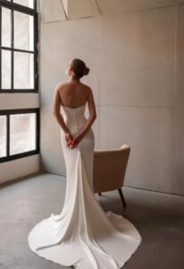 A bride shows the back of the Tommy by Neta Dover wedding dress with a fitted corset bodice and fabric-covered buttons on the back. The bridal gown features a full-length skirt with a side slit and is available in Chicago, IL.