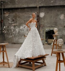 Woman standing in studio in a strapless gown featuring a sweetheart neckline and a voluminous circle skirt with a dramatic train, crafted from luxurious silk organza and adorned with delicate, handmade flower appliqués