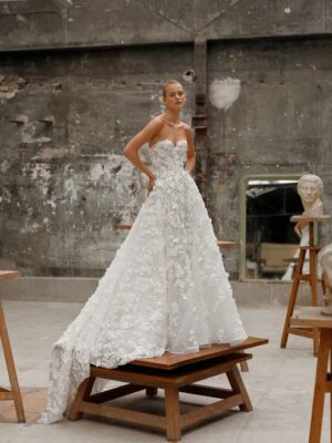 Woman standing in studio in a strapless gown featuring a sweetheart neckline and a voluminous circle skirt with a dramatic train, crafted from luxurious silk organza and adorned with delicate, handmade flower appliqués