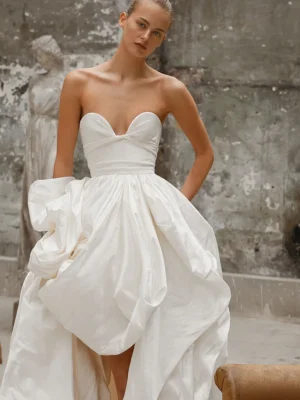 A bride wears Elle by Dana Harel, a strapless bridal gown elegantly fashioned from sumptuous taffeta silk that features a daring sweetheart neckline and a beautifully voluminous skirt, artfully hand-draped to cascade into a graceful train extension. See this wedding dress in Chicago, IL.