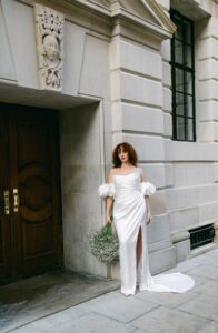 A bride wears a wedding dress, Tra Tra by Half Penny London. The bridal gown has a dreamy, soft-pointed tipped neckline emphasized by a draped liquid-like-satin corset flowing into a split skirt train. Available in Chicago, IL.