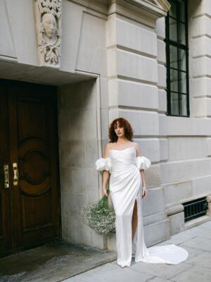 A bride wears a wedding dress, Tra Tra by Half Penny London. The bridal gown has a dreamy, soft-pointed tipped neckline emphasized by a draped liquid-like-satin corset flowing into a split skirt train. Available in Chicago, IL.