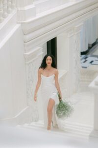 A bride walks upstairs wearing a wedding dress, Tra Tra by Half Penny London. The bridal gown has a dreamy, soft-pointed tipped neckline emphasized by a draped liquid-like-satin corset flowing into a split skirt train. Available in Chicago, IL.