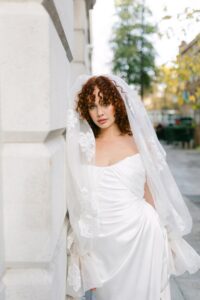 A close-up image of a bride wearing the wedding dress Tra Tra by Half Penny London. The bridal gown has a dreamy, soft-pointed tipped neckline emphasized by a draped liquid-like-satin corset flowing into a split skirt train. Available in Chicago, IL.