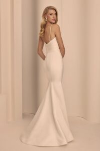 Sleeveless gown features a draped Duchesse Satin bodice, a V-neckline, and slender spaghetti straps. Fabric-covered buttons cascade down the back, ending in a flower embellishment at the waist. The draped skirt includes a slit, and a detachable skirt.